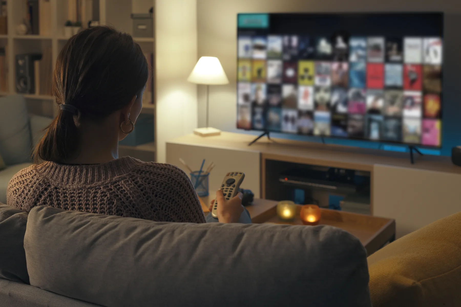 En person sitter i soffan i ett mysigt vardagsrum och tittar på tv med fjärrkontrollen i handen